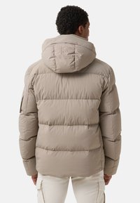 Man in een beige gewatteerde jas met capuchon en geborduurde 'B' op de achterkant, gecombineerd met witte cargobroek, gezien van achteren.