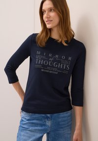 Marineblaue Langarm-T-Shirt mit Rundhalsausschnitt und grafischem Textdesign in hellen Farben. Kombiniert mit hellblauen Jeans.