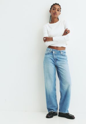 Svijetloplave široke traperice s izblijedjelim pranjem, uparene s krojenom bijelom bluzom s dugim rukavima i crnim cipelama, na jednostavnoj bijeloj pozadini.