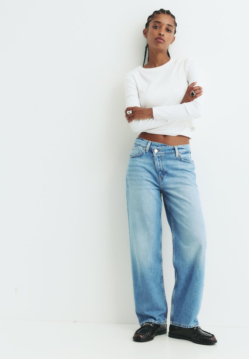 Svijetloplave široke traperice s izblijedjelim pranjem, uparene s krojenom bijelom bluzom s dugim rukavima i crnim cipelama, na jednostavnoj bijeloj pozadini.