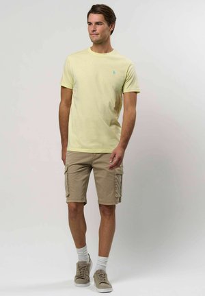 Mladic nosi svijetložutu majicu, bež kratke hlače, bijele čarape i bež tenisice, stojeći protiv jednostavne svijetle pozadine.