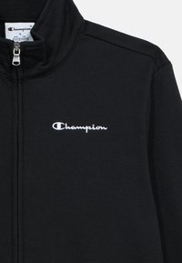 Μαύρο μπουφάν με φερμουάρ, κατασκευασμένο από απαλό ύφασμα, με όρθιο γιακά και κεντημένο λευκό λογότυπο "Champion" στο αριστερό μέρος του στήθους.