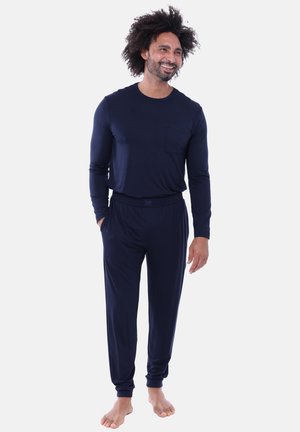Man met krullend haar en baard, gekleed in een donkerblauw shirt met lange mouwen en bijpassende broek, staat blootsvoets op een witte achtergrond.