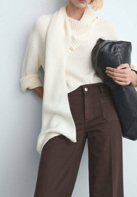 Persona con un suéter acanalado color crema y pantalones marrón oscuro, sosteniendo un bolso grande de cuero negro sobre un fondo liso y claro.