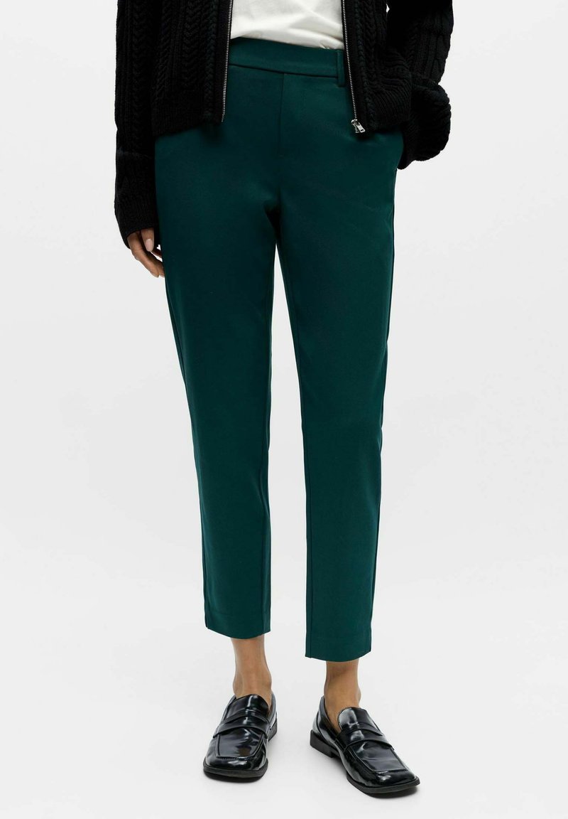 Teal tailored trousers with a smooth texture, featuring a straight cut and ankle length, paired with black patent loafers.