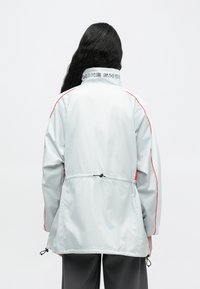 Helehall tuulekindel jope punaste detailidega, reguleeritava paelaga vöökohal ja kaelusel oleva "NIKE SHOE" logoga.