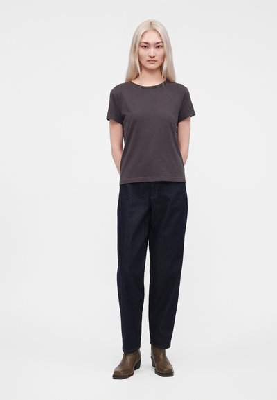 Femme aux longs cheveux blonds debout, portant un t-shirt gris foncé, un jean bleu foncé et des bottines marron sur un fond clair uni.