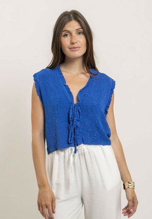 Femme aux longs cheveux bruns portant un haut bleu texturé sans manches avec des liens à l'avant et un pantalon blanc ample, debout devant un fond uni.