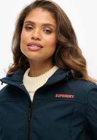 Granatowa kurtka soft-shell z wysokim kołnierzem, zamkiem błyskawicznym i haftowanym pomarańczowym logo "SUPERDRY". Pod spodem biały sweter z prążkowanym wzorem.