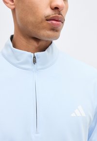 Κοντινό πλάνο ενός άνδρα που φοράει αθλητική μπλούζα ανοιχτού γαλάζιου με φερμουάρ στο ένα τέταρτο και λευκό λογότυπο Adidas στο στήθος.