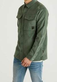 Vihreä samettipaitapaita, jossa on napillinen edusta, kaksi rintataskua ja teksturoidun pinnan viimeistely. Vasemman rintataskun päällä on logopainatus.