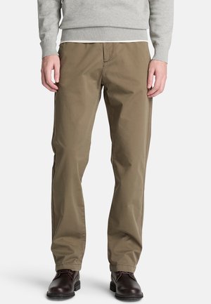 Hombre con pantalones marrones de corte recto, zapatos de vestir marrón oscuro y un suéter gris, de pie frente a un fondo blanco liso.