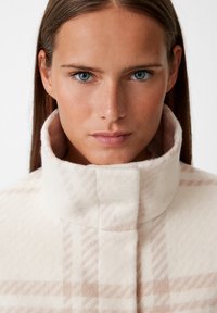 Cappotto a scacchi crema e rosa con alto colletto; tessuto morbido e testurizzato; chiusura a bottone al collo; design minimalista, senza hardware visibile.