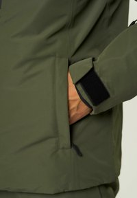 Oliiviroheline jakk sujuva, veekindla kangaga. Omab külgprahti ja Velcro mansette, et reguleerida sobivust.