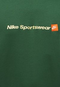 Πράσινο ύφασμα με κρεμ κείμενο "Nike Sportswear" και πορτοκαλί τετράγωνο μπαλώμα με λευκό λογότυπο Nike swoosh.