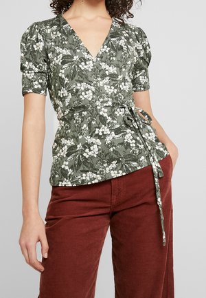 Vrouw draagt een groene bloemen overslagblouse met pofmouwen en roestkleurige ribfluwelen broek, hand in de zak, tegen een neutrale achtergrond.