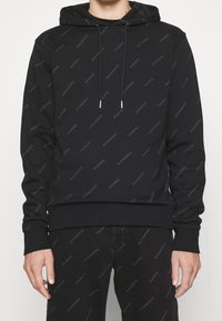 Sudadera negra con un patrón de logotipos repetido, capucha con cordón, puños y dobladillo acanalados, y textura de tela suave. Estilizada con jeans negros.