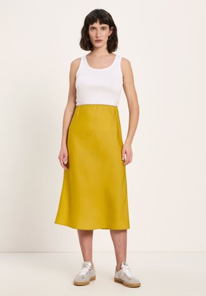 Woman with short dark hair wearing a white sleeveless top, mustard midi skirt, and white sneakers standing against a plain background.