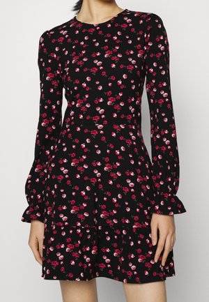 Femme portant une robe noire à manches longues avec un motif floral de roses rouges et roses et une jupe légèrement évasée.