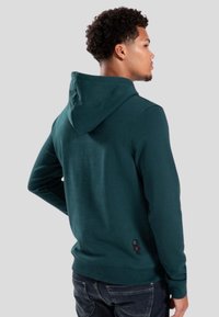 Felpa con cappuccio verde scuro dalla finitura testurizzata, dotata di cappuccio regolabile con lacci e un piccolo dettaglio del personaggio ricamato vicino alla parte inferiore.