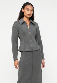 Sivi zip-up jakna s peplum dizajnom, s oštrim ovratnikom i dugim rukavima, izrađena od mekog, teksturiranog materijala.