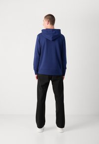 Sudadera azul marino con capucha ajustable, hecha de material suave, combinada con pantalones negros y zapatillas blancas, vista desde la parte de atrás.