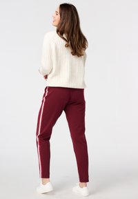 Pantalons de survêtement bordeaux avec des rayures blanches sur les côtés, associés à un pull crème à côtes. Le mannequin se tient dans une posture détendue. Baskets blanches visibles.