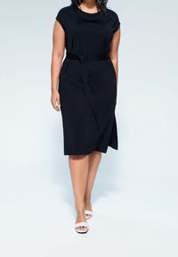 Schwarzes Kleid mit kurzen Ärmeln, tailliertem Bund und asymmetrischem Saum. Tragen mit transparenten, hochhackigen Sandalen. Glatte Stoffoberfläche.