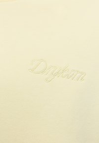 Tissu de couleur crème avec un texte cursif brodé discret "Drykorn" en fil assorti.