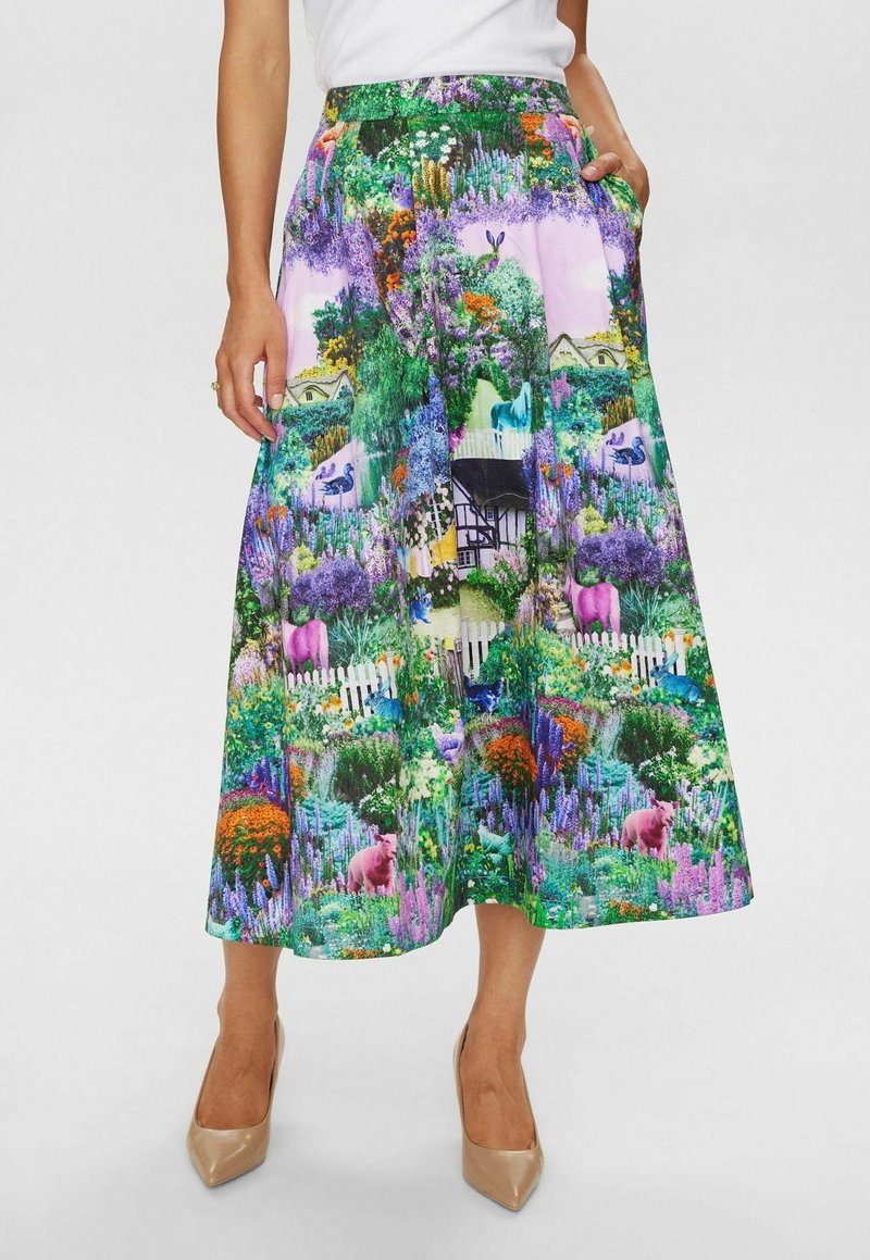Femme portant une jupe midi colorée avec un imprimé détaillé de jardin et d’animaux, associée à des escarpins beiges à bout pointu et un haut blanc.