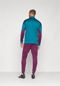 Teal ja maroon värvi track-jakk, millel on pikad varrukad, koos vastava maroon värvi pükstega. Valmistatud siledast kangast, millel on elastsed mansetid.