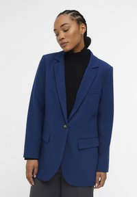 Blauwe blazer van textuurstof, met een notch-lapel, een enkele knoopsluiting en voorzakken. Draag het over een zwarte top.