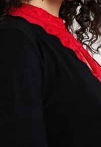 Schwarzer Pullover mit einem roten Spitzensaum, mit rundem Ausschnitt und strukturiertem Stoff. Nahaufnahme des Ausschnitts.