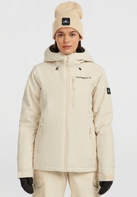 Beige waterdichte jas met een capuchon, ritszakken en een logo op de borst; gedragen met een bijpassende beige beanie en zwarte handschoenen.