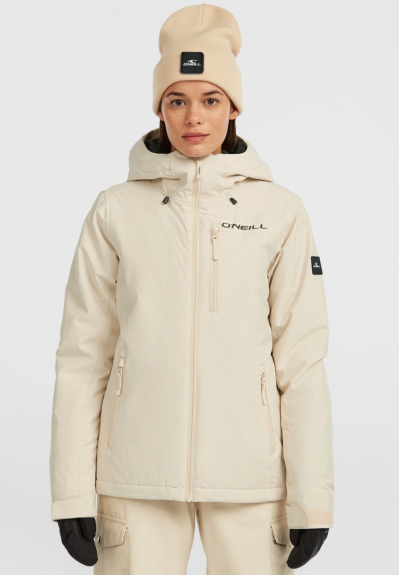 Beige waterdichte jas met een capuchon, ritszakken en een logo op de borst; gedragen met een bijpassende beige beanie en zwarte handschoenen.