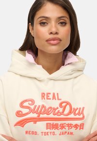 Crème hoodie met een prominent rood-oranje "SuperDry" logo en Japanse karakters, met een zachte textuur en een capuchon met trekkoord.