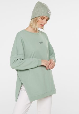 Woman wearing a sage green oversized sweatshirt and matching knit beanie, looking down with hands gently clasped.