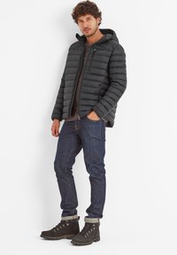 Black puffer jacket with horizontal quilting, zippered chest pocket, and hood, worn over a dark shirt and blue jeans with roll-up cuffs. Brown hiking boots.