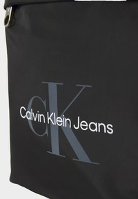 Borsa in tessuto nero con la scritta "Calvin Klein Jeans" in bianco e grigio. Il design presenta un logo prominente e un'apertura con zip.
