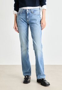 Jean bleu clair avec une coupe droite, en denim. Comprend un design classique à cinq poches et une légère décoloration. Porté avec des chaussures noires.