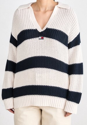 Personne portant un pull à col en V à maille lâche avec de larges rayures horizontales bleu marine et blanc et un petit logo Tommy Jeans sur la poitrine.