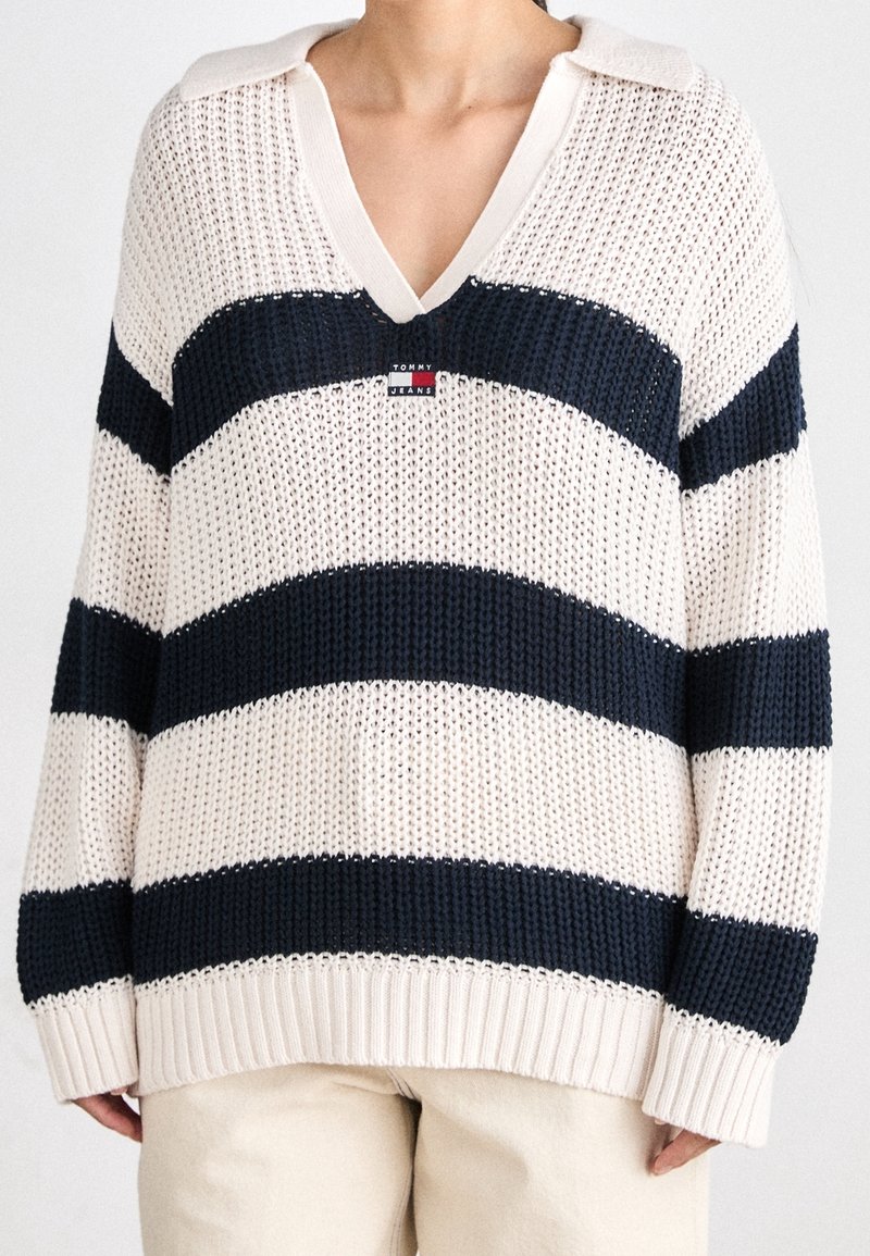 Personne portant un pull à col en V à maille lâche avec de larges rayures horizontales bleu marine et blanc et un petit logo Tommy Jeans sur la poitrine.