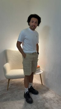 Άνδρας με αφρο-χτένισμα που φοράει λευκό μπλουζάκι, μπεζ cargo σορτς, λευκές κάλτσες και μαύρα αθλητικά παπούτσια, στέκεται δίπλα σε μια λευκή υφασμάτινη πολυθρόνα και ένα τραπέζι.