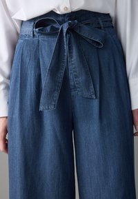 Pantalones de mezclilla de corte amplio en azul oscuro con una cintura alta, con un cinturón anudado en la parte delantera y una textura sutil. Combinados con una camisa blanca.