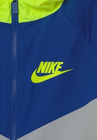 Giacca Nike caratterizzata da un corpo blu con cappuccio, zip e logo giallo neon. Il design include un motivo geometrico con accenti grigi.