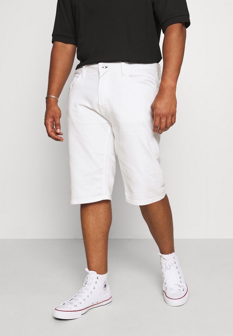 Homme portant un short blanc aux genoux, des baskets blanches montantes et une chemise noire à manches courtes, debout sur un sol gris clair.