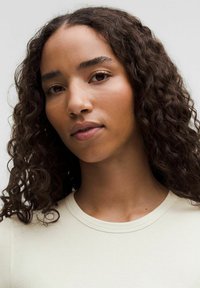 Svetlo béžové rebrované tričko s okrúhlym výstrihom, ktoré nosí osoba s kučeravými vlasmi a prirodzeným make-upom, ukazujúc hladkú pokožku a neutrálnymi tónmi.