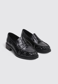 Mocasines de cuero negro con un acabado texturizado, que presentan un borde acanalado y suela plana. Diseño moderno con sutiles detalles de costura.