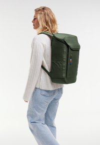 Sac à dos vert olive avec une forme structurée, doté d'une fermeture à rabat, d'une poche frontale avec fermeture éclair et de bretelles ajustables. Texture de tissu lisse.