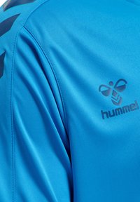 Jasny niebieski materiał sportowej koszulki z widocznym szwem na ramieniu oraz czarnym logo „hummel” z stylizowanym emblematem pszczoły na piersi.